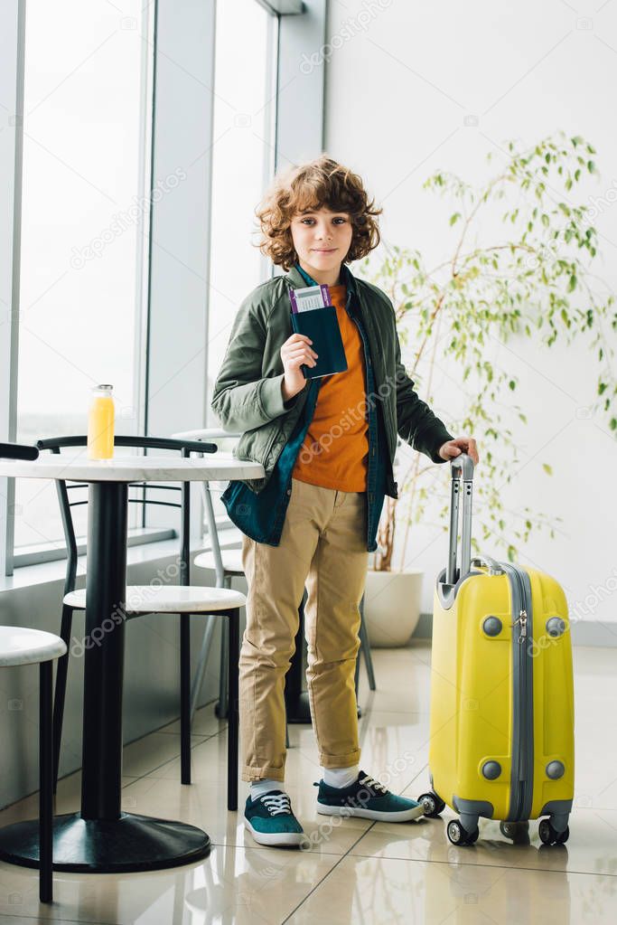 full length view of boy standing with yellow suitcase, holding passport and ticket in waiting hall in airport