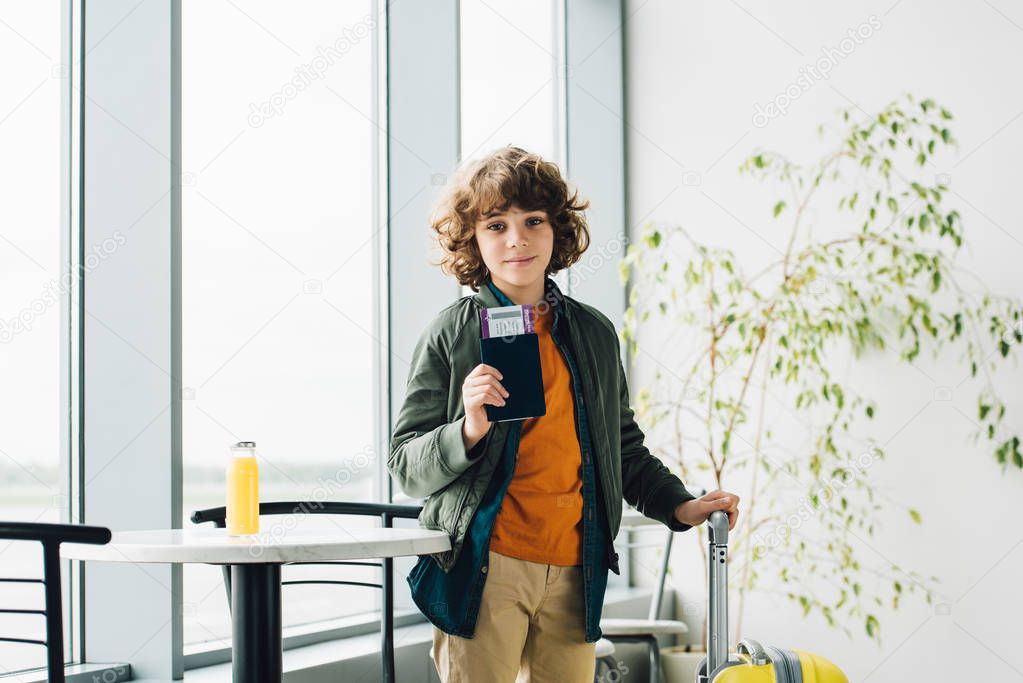 boy standing with yellow suitcase, holding passport and ticket in airport 