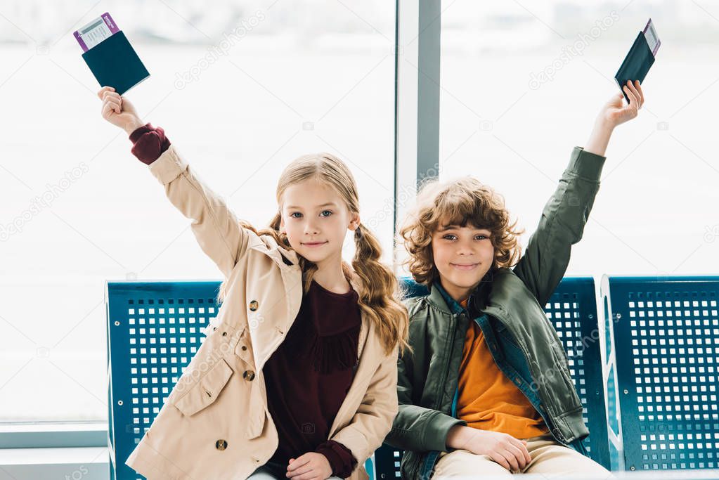 happy kids sitting on blue seats, holding passports and tickets in waiting hall in airport