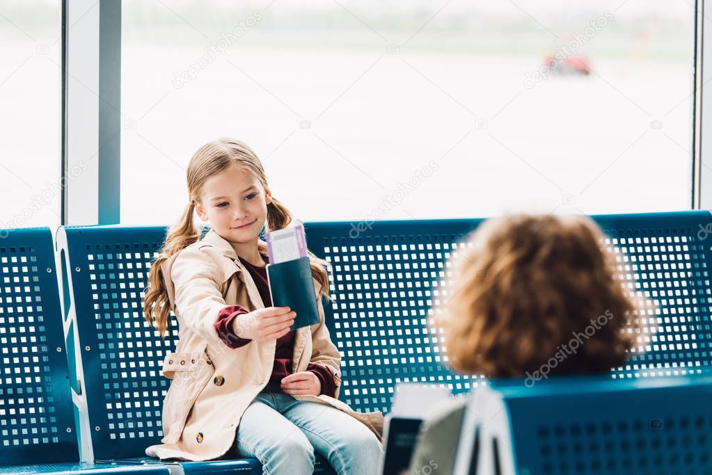 preteen kid sitting on blue seat, showing passport to boy in waiting hall in airport 