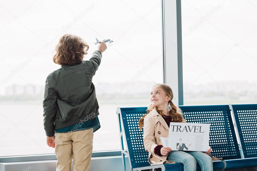cute preteen kid sitting in waiting hall with travel newspaper while boy playing with toy plane