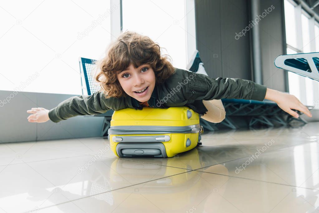 excited preteen kid lying on suitcase with outstretched hands in airport departure lounge
