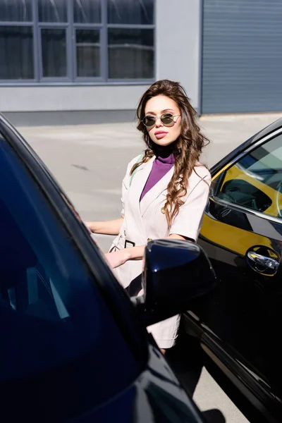 selective focus of serious young woman in sunglasses standing near cars