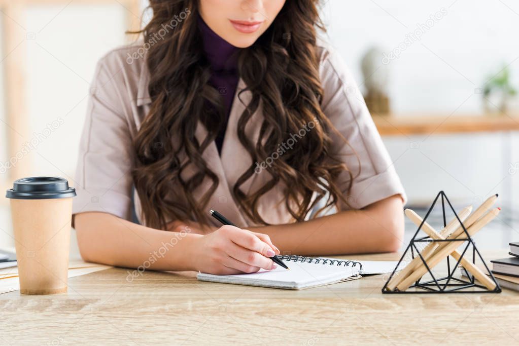 cropped view of curly girl writing in notebook near paper cup 