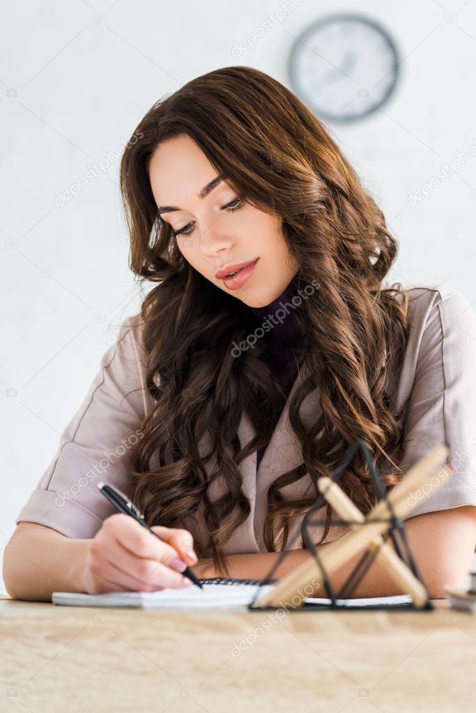 selective focus of attractive curly girl writing in notebook near stationary 