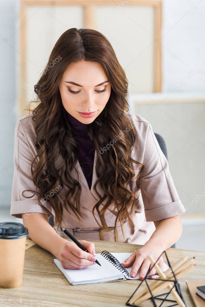 selective focus of pretty curly girl writing in notebook near stationary and paper cup