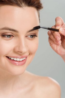gri izole neşeli çıplak kadın yakınında kaş fırçası tutan makyaj sanatçısı kırpılmış görünümü 