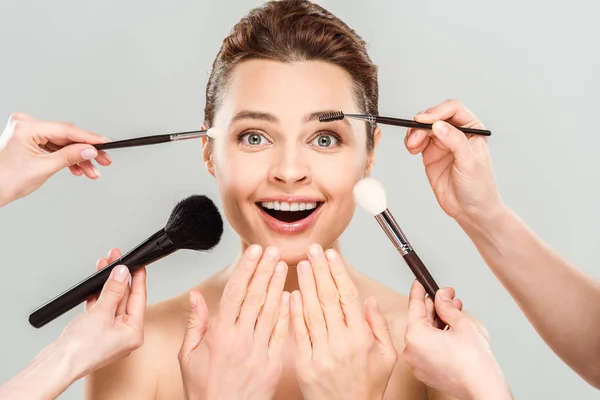 stock image cropped view of makeup artists holding cosmetic brushes near naked excited woman isolated on grey 