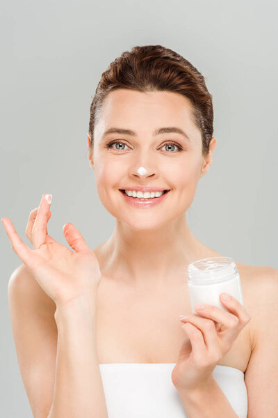 cheerful woman applying face cream and smiling isolated on grey 