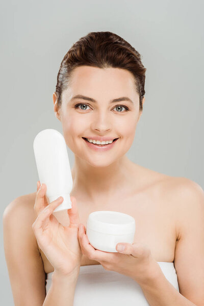 cheerful woman holding container with cosmetic cream and bottle of body lotion isolated on grey 