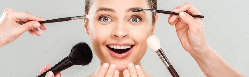 Panoramic shot of makeup artists holding cosmetic brushes near excited woman isolated on grey — Stock Image Panoramic Shot Makeup Artists Holding Cosmetic Brushes Excited Woman Isolated Stock Image
