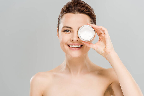 cheerful naked woman covering eye while holding container with face cream near eye and smiling isolated on grey 