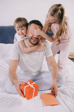 happy man holding gift box and fathers day greeting card while adorable kids hugging him and covering his eyes with hands clipart
