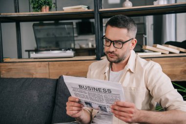 handsome man in glasses reading business newspaper while sitting on sofa at home clipart