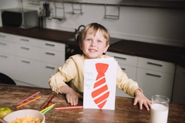 cute boy showing fathers day greeting card with drawn tie while sitting at kitchen table and smiling at camera clipart
