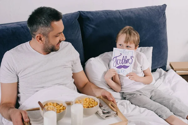 お父さんの日のグリーティングカードで顔を覆うかわいい男の子は トレイに朝食とパパの近くのベッドに座っている間 — ストック写真