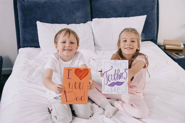 adorable children holding fathers day greeting cards while sitting on bedding and smiling at camera