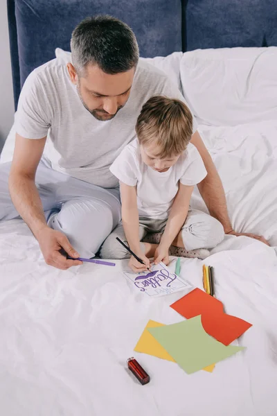 cute son drawing fathers day greeting card while sitting on bedding near daddy