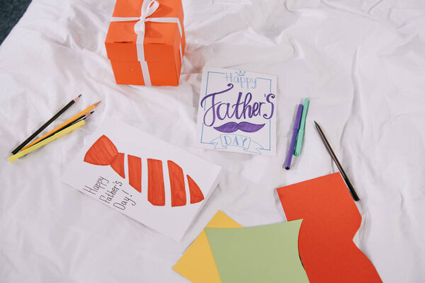 happy fathers day greeting card with drawn mustache and tie, gift box, pencils, felt pens and multicolored sheets of paper on white bedding
