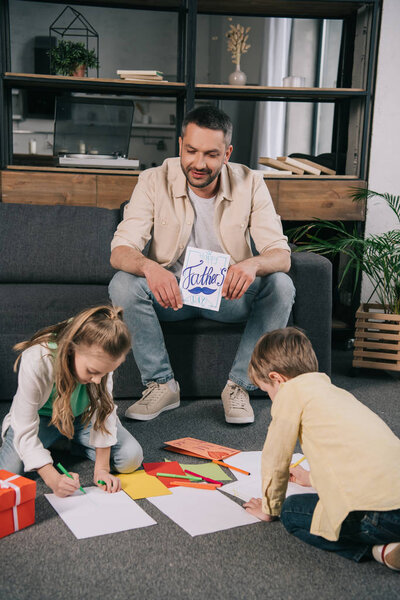 happy man sitting on sofa and holding fathers day greeting card near children drawing on while paper sheets while sitting on floor