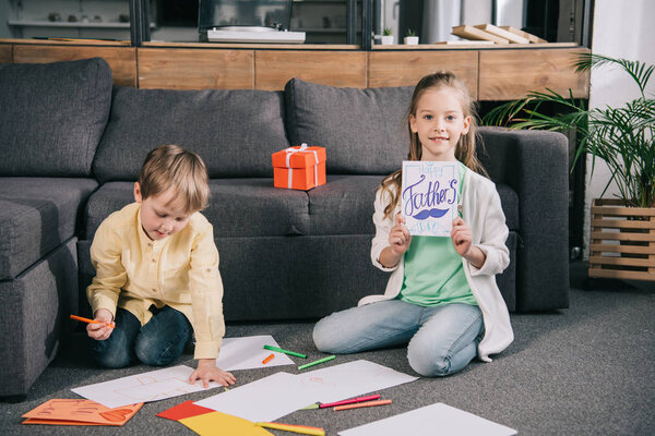 cute child showing fathers day greeting card while sitting on floor near brother drawing on white paper sheets