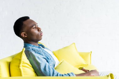side view of calm african american man sitting on couch at home clipart