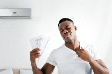 sweaty african american man closing eyes, standing in white room with heat temperature, holding newspaper in hand clipart