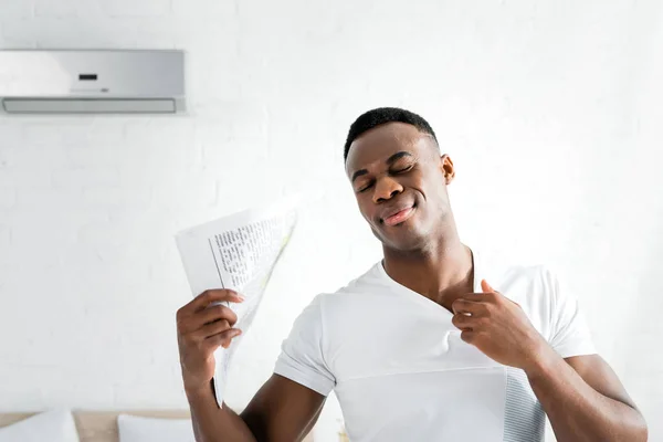 汗をかいたアフリカ系アメリカ人男性が目を閉じ 暑さの白い部屋に立ち 新聞を手に持つ — ストック写真