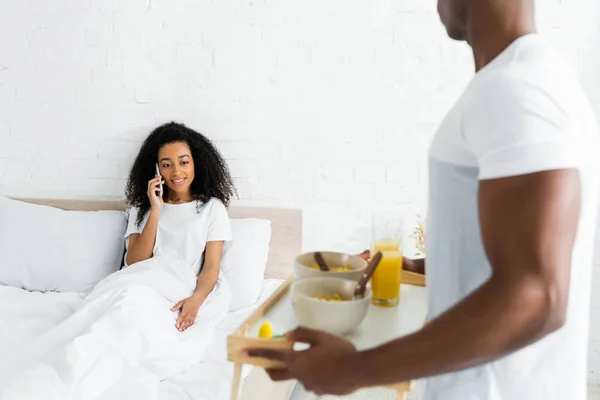 选择性的焦点 非洲美国女人在智能手机上说话 而男友拿着托盘与早餐 — 图库照片