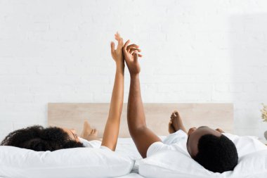 happy african american man touching hands with girlfriend, lying on white bed in bedroom clipart