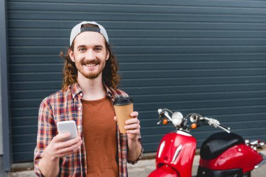 Elinde kahve ve akıllı telefonla kırmızı scooter 'ın yanında duran kağıt bardak tutan genç adam. 