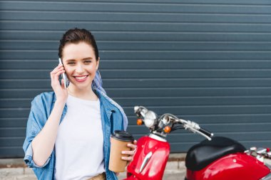 kırmızı scooter yanında duran güzel kız ön görünümü, kahve ile kağıt fincan tutan ve akıllı telefon konuşurken