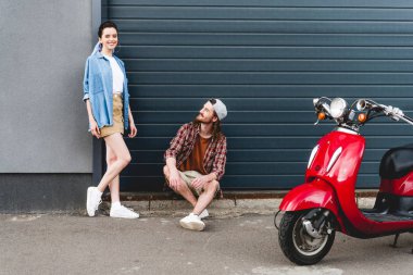 beautiful girl standing near wall and young man sitting on the floor near red scooter and looking at girl  clipart
