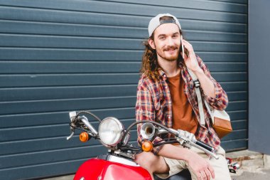 Genç yakışıklı adam kırmızı scooter 'da oturuyor, akıllı telefonla konuşuyor ve kameraya bakıyor. 