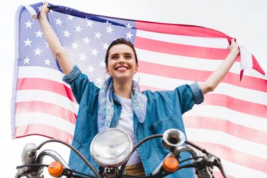 scooter üzerinde oturan güzel kız düşük açı görünümü, Amerikan bayrağı tutan ve gökyüzü arka planda gülümseyen