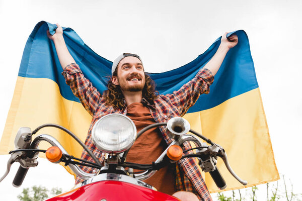 low angle view of young man sitting on red scooter and holding Ukrainian flag 
