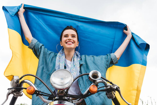 низкий угол обзора красивой девушки, сидящей на скутере, улыбающейся и держащей украинский флаг
