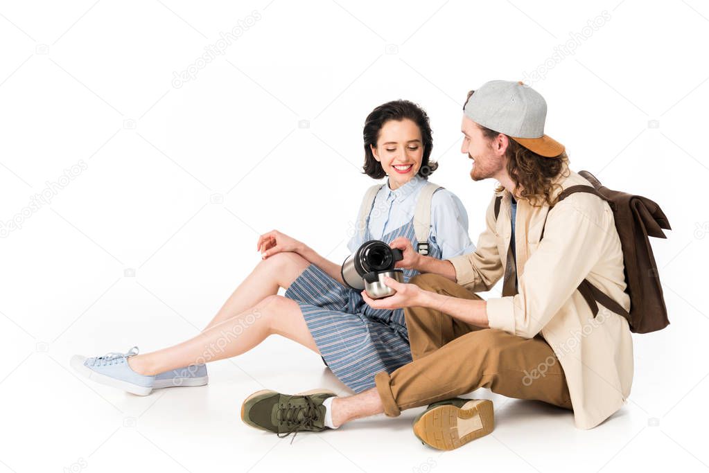 young couple of tourists sitting and man pouring cup from thermos isolated on white