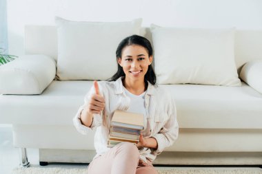 evde yerde otururken kitap tutan ve başparmak gösteren mutlu latin kadın