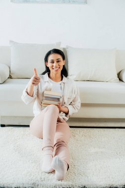 neşeli latin kadın başparmak gösteren ve evde yerde otururken kitap yığını tutarak