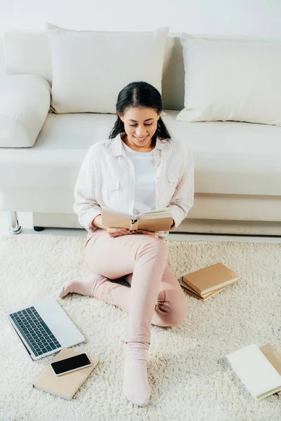 Leende Latin Kvinna Läsa Bok När Sitter Golvet Nära Laptop — Stockfoto