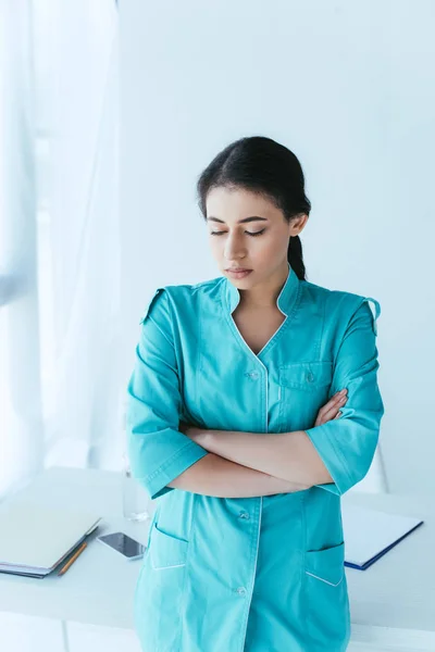 offended latin doctor standing at workplace with crossed arms and looking down