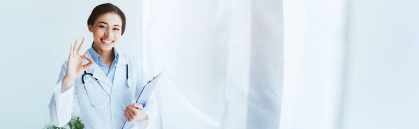 panoramic shot of happy latin doctor showing ok gesture and holding clipboard