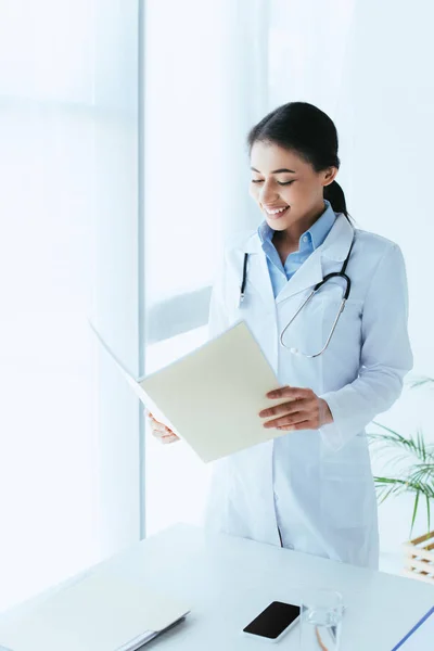 病院で窓のそばに立っている間 紙のフォルダを保持している笑顔のラテン医師 — ストック写真