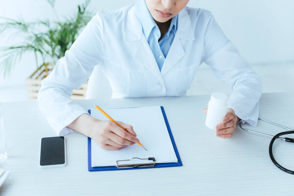 cropped shot of young doctor writing on clipboard while sitting at workplace and holding pills container