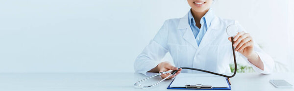 panoramic shot of smiling doctor holding stethoscope near clipboard