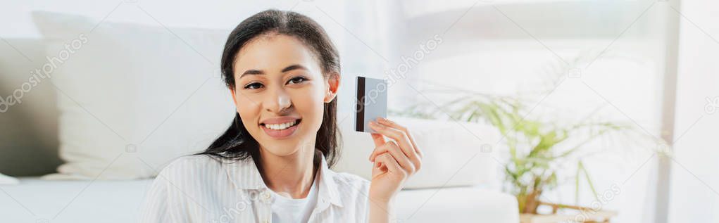 panoramic shot of cheerful latin woman holding credit card and looking at camera