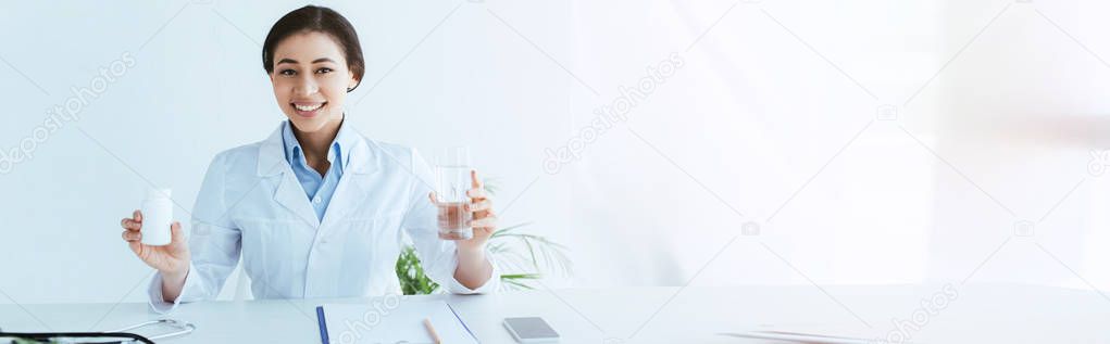 panoramic shot of smiling latin doctor holding glass of water and container with pills 