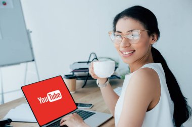 Kiev, Ukrayna - 26 Nisan 2019: Ekranda Youtube web sitesi ile dizüstü bilgisayar kullanırken kahve fincanı tutan çekici latin iş kadını.