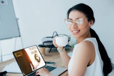ekranda bilet online web sitesi ile dizüstü bilgisayar kullanırken kahve fincanı tutan güzel latin iş kadını